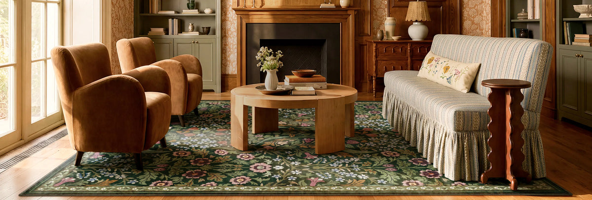 A styled living room featuring a large green and blue floral area rug under a round wooden coffee table, flanked by two brown velvet armchairs on one side and a cream striped sofa on the other.
