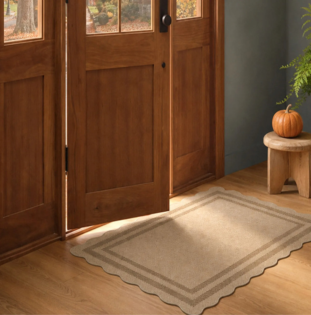 A rectangular entryway rug with a scalloped border in beige and brown tones, placed on a wood floor in front of open wooden double doors in a home entryway.