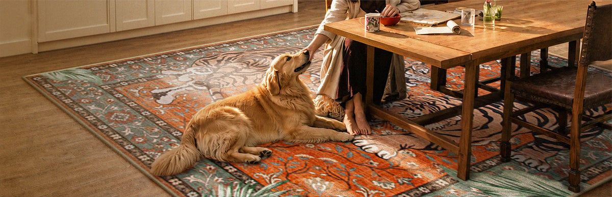 A large traditional-style area rug with a red, blue, and beige floral pattern is shown in a dining room setting, featuring a woman sitting at a wooden table and a golden retriever lying on the rug.